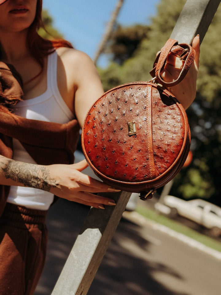 Bolsa redonda Strut em couro de avestruz cheio caramelo segurada na mão, textura dos folículos visível